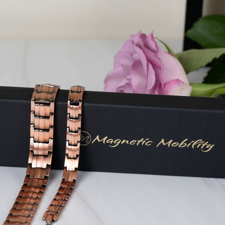 Ashe Copper Magnetic Bracelets - 9mm slim band and 15mm wide band - displayed draped over a Magnetic Mobility gift box with a pink rose in the background.