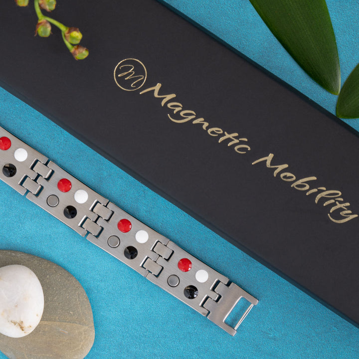 The reverse side of the Magnetic Mobility Yarrow Star 4in1 Magnetic Bracelet is featured, displaying a double row of therapeutic elements including Neodymium magnets, Germanium inserts, Negative ions, and FIR elements. The detailed view against a blue backdrop highlights the health-focused design, with the brand's sophisticated black box and a natural green element adding to the aesthetic appeal.