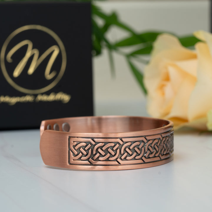 Side view of the Burdock Copper Bracelet featuring intricate Celtic knot detailing, displayed alongside a Magnetic Mobility box and a yellow rose.