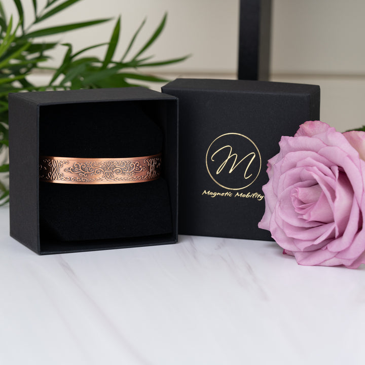 Campion Copper Bracelet displayed elegantly in its presentation box, highlighting its luxurious packaging and engraved floral design. The black Magnetic Mobility logo and a rose enhance the visual appeal.