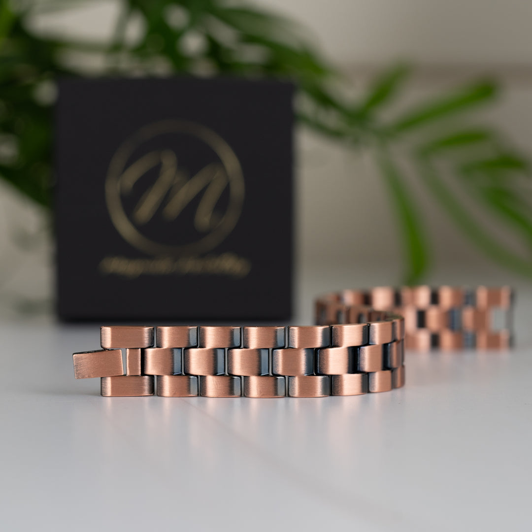 Front view of the Columbine Copper Bracelet in a slight twist, emphasising its classic link design and polished finish. The Magnetic Mobility gift box and a green plant are visible in the background.