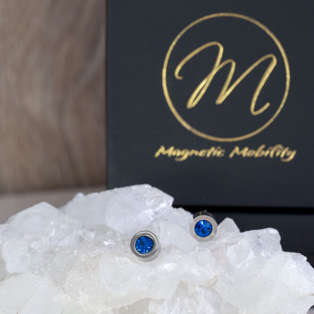 Pair of December birthstone magnetic stud earrings with vibrant Swarovski Blue Zircon, shown from the front view against a backdrop of clear crystals and Magnetic Mobility packaging.