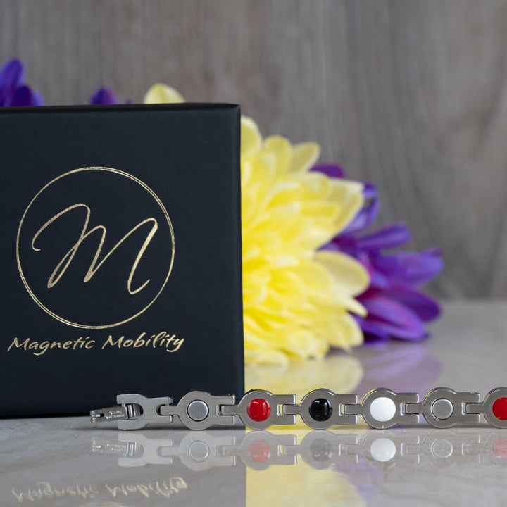 View of the back of a 4in1 Magnetic bracelet showing the 4 elements, magnets, FIR element, Germanimum and Negative ions. There is a black gift box in the background and yellow and purple flowers.