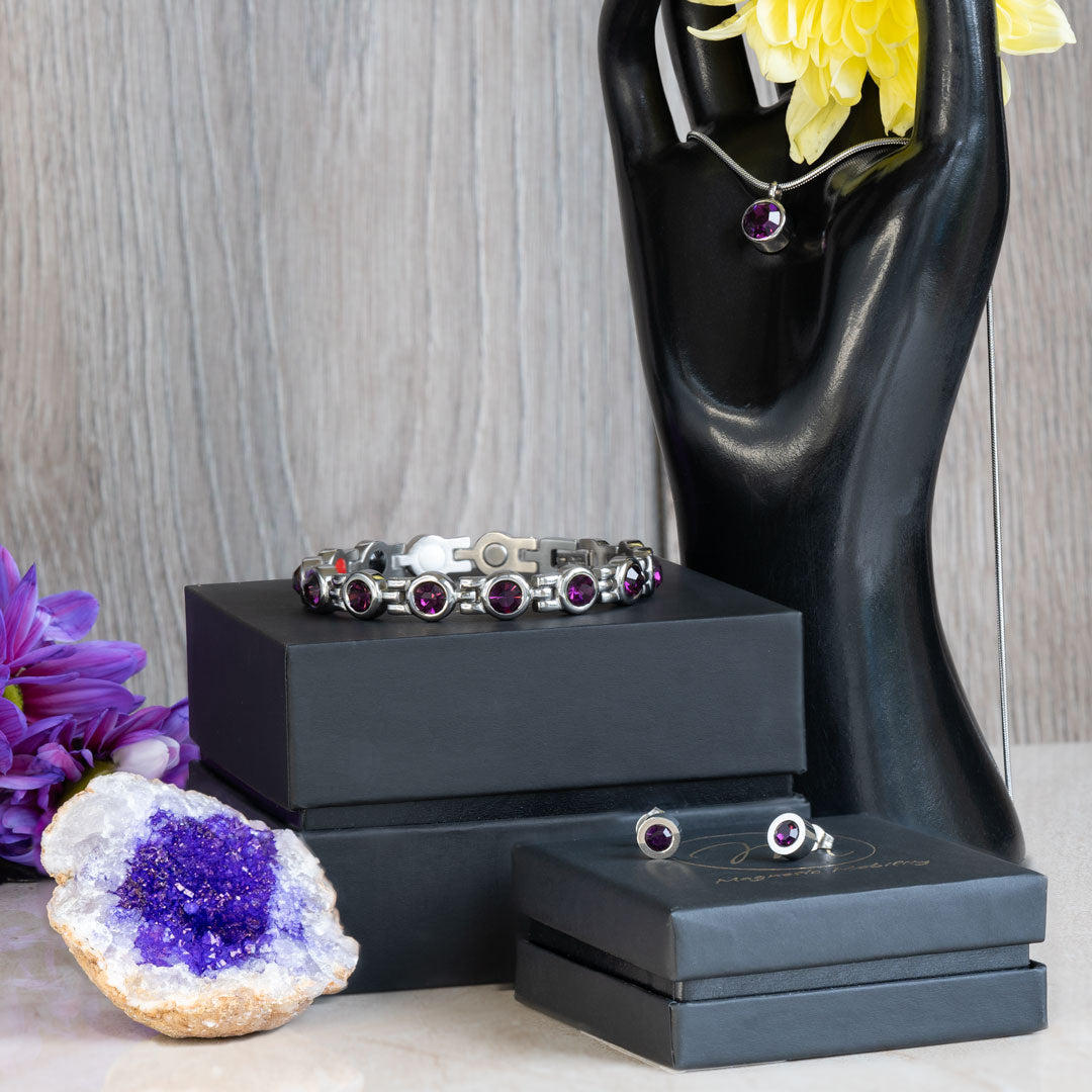 Febraury Birthstone gift set containing a 4in1 Magnetic bracelet with February birthstone Swarovski Amethyst crystals, a February birthstone necklace with one Swarovski Amethyst crystal and a neo-dynimum magnet in the back and a pair of February Birthstone magnetic earrings with Neodynimum magnets in the back. The necklace is on a black stand in the shape of a hand, the earrings are on a gift box. There is an amethyst geode in the middleground.