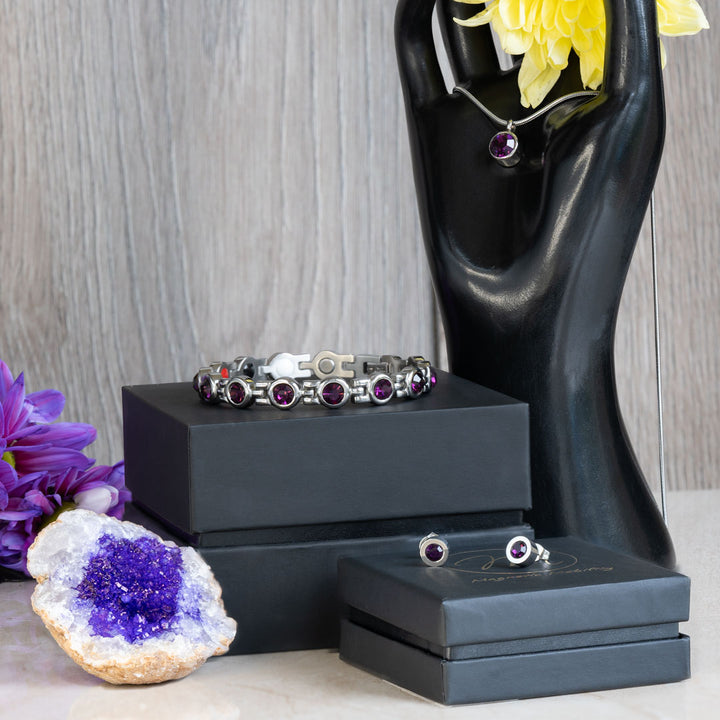 Febraury Birthstone gift set containing a 4in1 Magnetic bracelet with February birthstone Swarovski Amethyst crystals, a February birthstone necklace with one Swarovski Amethyst crystal and a neo-dynimum magnet in the back and a pair of February Birthstone magnetic earrings with Neodynimum magnets in the back. The necklace is on a black stand in the shape of a hand, the earrings are on a gift box. There is an amethyst geode in the middleground.
