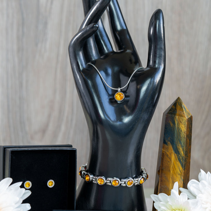 The complete November Birthstone Gift Set, including a citrine magnetic necklace, earrings, and bracelet, is showcased in an elegant display with a dark hand model and flowers, emphasizing the bright, sunny yellow of the citrine stones.