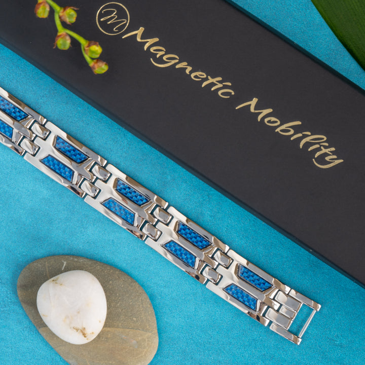 Top view of the Magnetic Mobility Yarrow Star 4in1 Magnetic Bracelet elegantly placed on a blue surface, showcasing its silver link design with vibrant blue carbon fiber inlays. The brand's sophisticated black box with gold script is partially visible, adding to the premium presentation of this health-conscious accessory, perfect for Google Shopping viewers seeking style and wellness.
