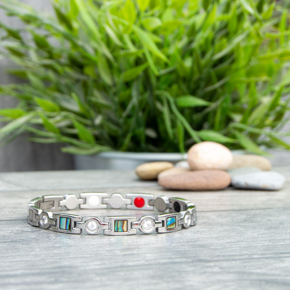 45 degree view of Avens Moon Womens Silver Magnetic Bracelet with Irredessant desings and white srystals. Image shows the bracelet on a wooden surface with green plants and pebbles.