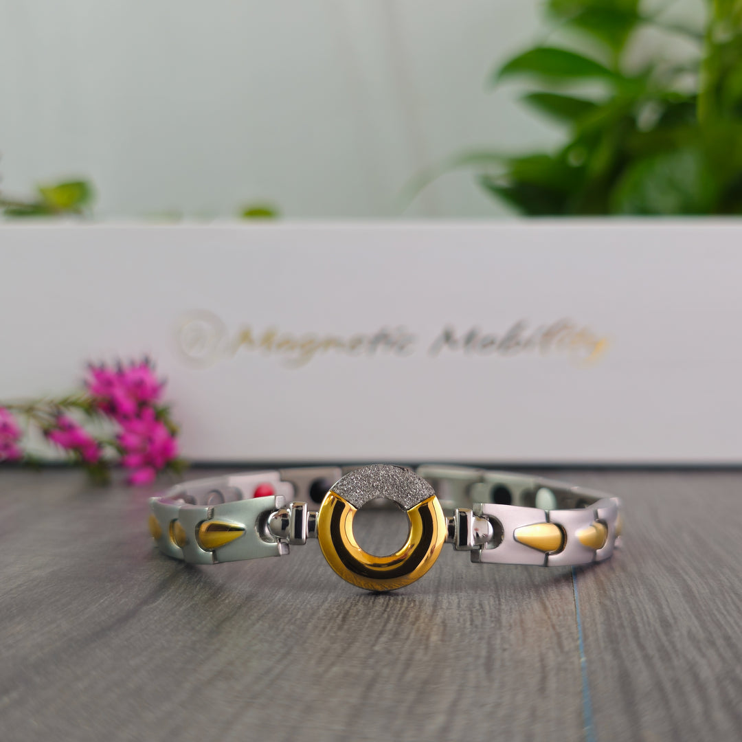 The Sorrel Full Moon 4in1 Magnetic Bracelet from Magnetic Mobility is displayed prominently on a grey surface. The bracelet features a striking combination of silver and gold segments and a central sparkling piece, with the brand's white box and a spray of pink flowers softly focused in the background.