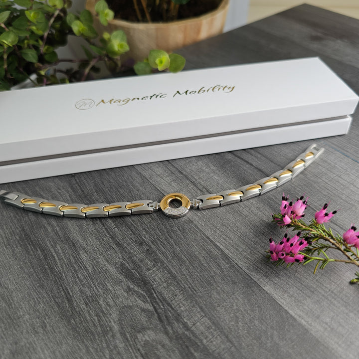 A Sorrel Full Moon 4in1 Magnetic Bracelet from Magnetic Mobility lies against a grey wooden surface with a branded white box in the background. The bracelet features alternating silver and gold links and a central circular piece with a sparkling stone, complemented by delicate pink flowers to the side