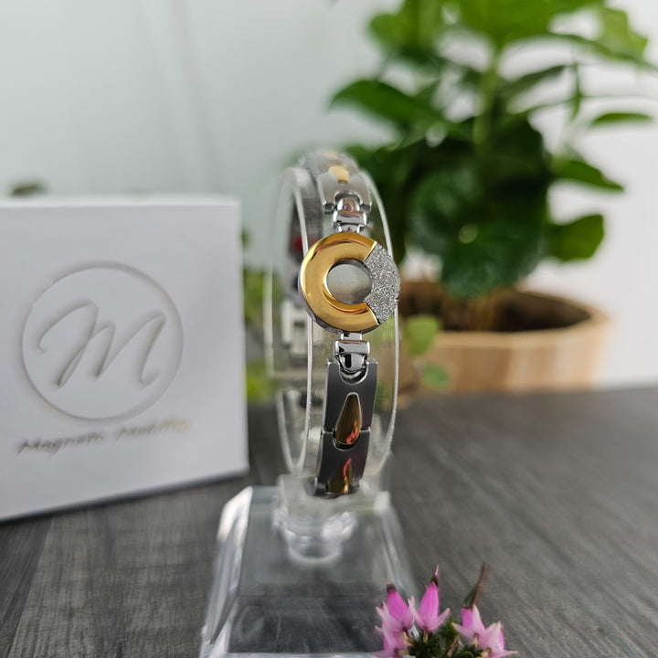 An elegant Sorrel Full Moon 4in1 Magnetic Bracelet from Magnetic Mobility is displayed on a clear stand with a lush green plant in the background, emphasizing the bracelet's silver and gold tones and the sparkling accent on the focal center