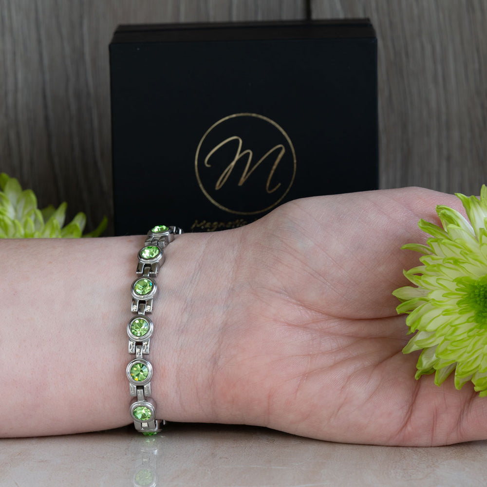 Close-up of the August Birthstone 4in1 magnetic bracelet adorned with Swarovski Peridot crystals, shown on a wrist with green floral elements and a black gift box in the background. This bracelet combines vibrant beauty and therapeutic benefits, making it a perfect gift for August.