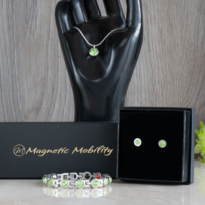 Elegant display of the August Birthstone magnetic gift set featuring a necklace, earrings, and bracelet with Swarovski Peridot crystals, set against a black backdrop and complemented by green flowers. Ideal for celebrating August birthdays with a touch of sophistication and health benefits.