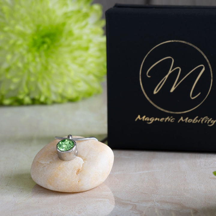 The August Birthstone magnetic necklace featuring a striking Swarovski Peridot pendant, elegantly displayed on a stone with a black gift box and green floral backdrop. Perfect for adding a touch of August elegance and wellness to any outfit.