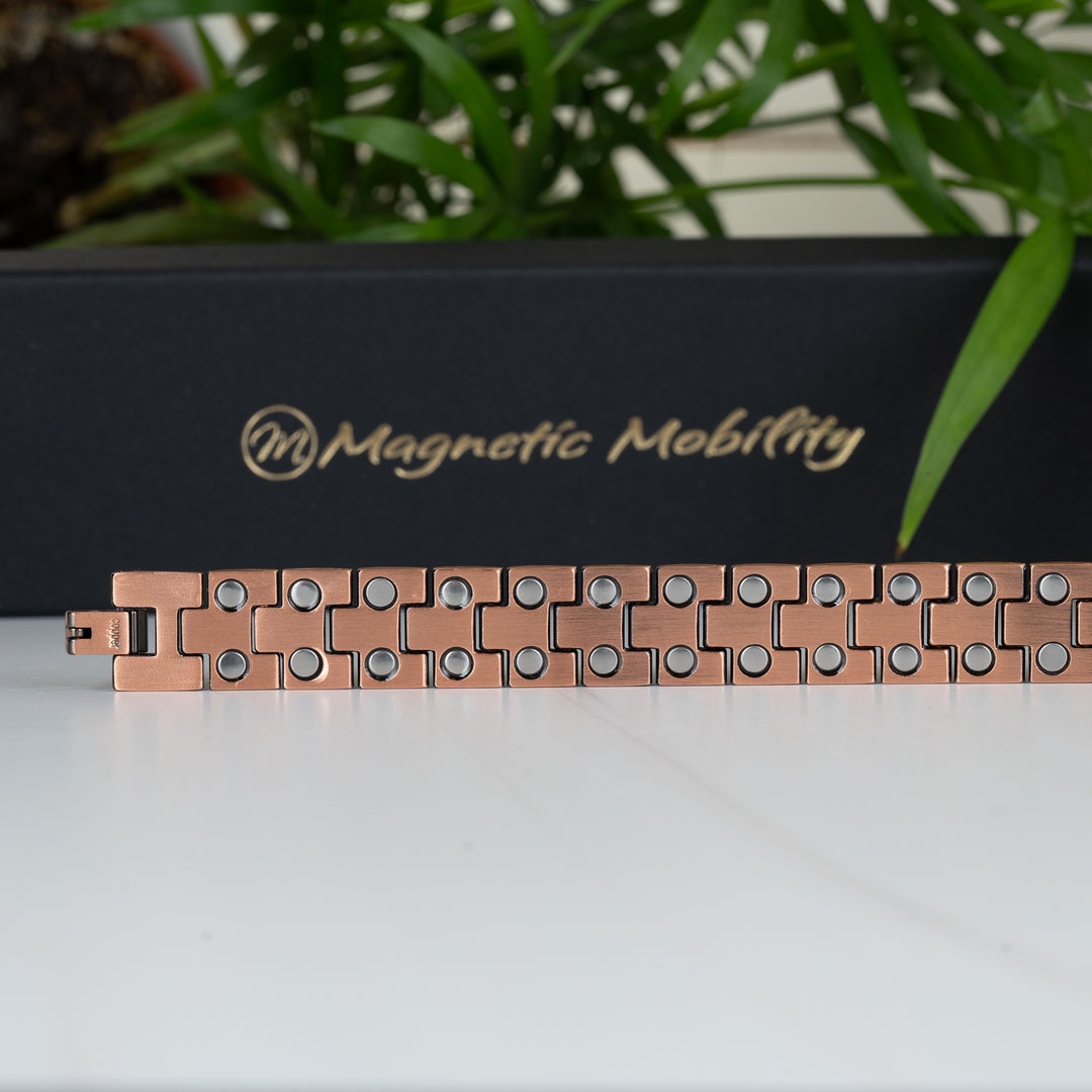 Close-up of the Columbine Copper Bracelet lying flat, showcasing the interior side with multiple double-strength neodymium magnets on each link. Black gift box with Magnetic Mobility logo in the background.