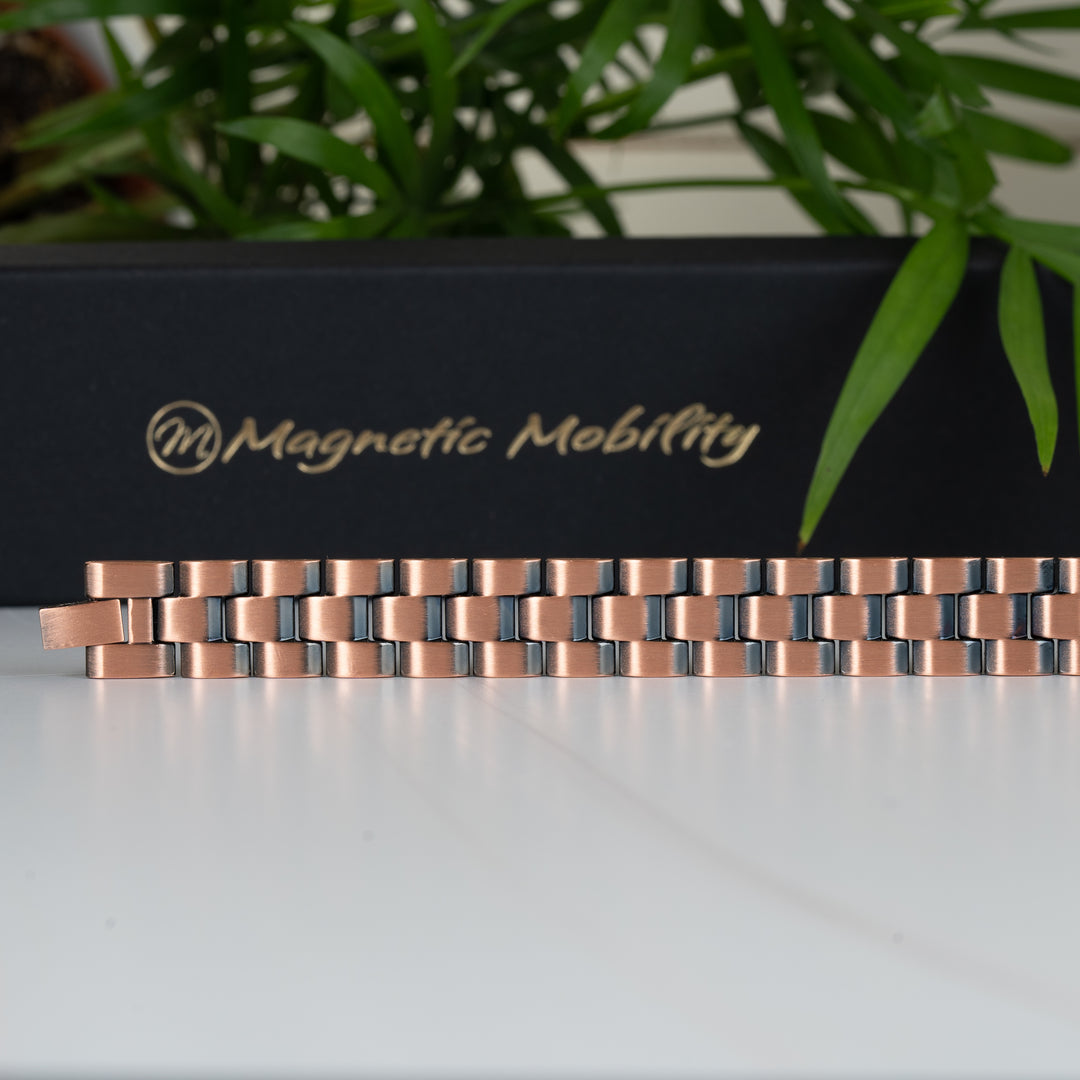 Full view of the Columbine Copper Bracelet, laid out flat to show the entire length and symmetrical link pattern. The backdrop features a green plant and the Magnetic Mobility logo.