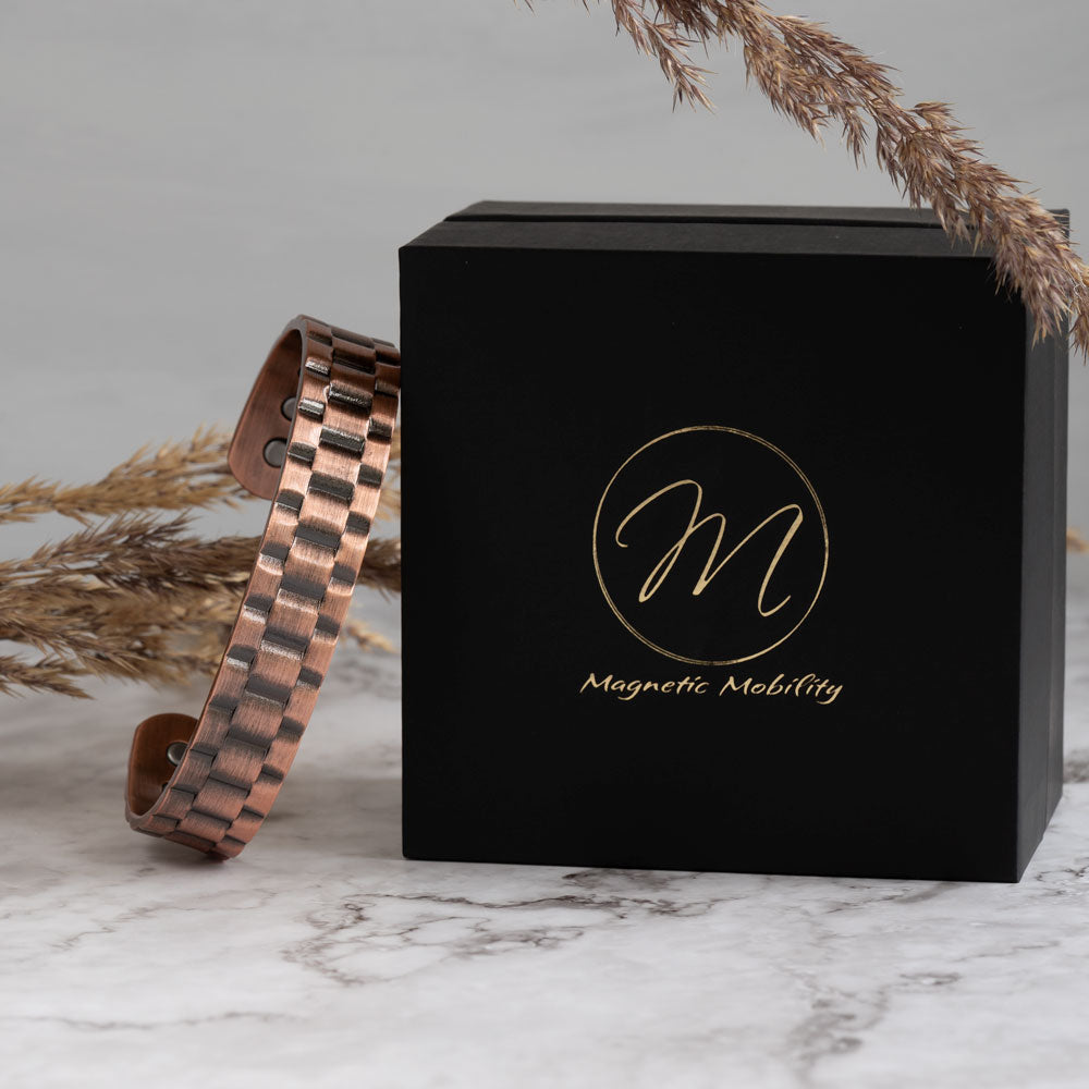 Elegant Columbine Copper Bracelet with detailed band design, leaning against a black Magnetic Mobility box, set on a marble surface with decorative dried grass accent