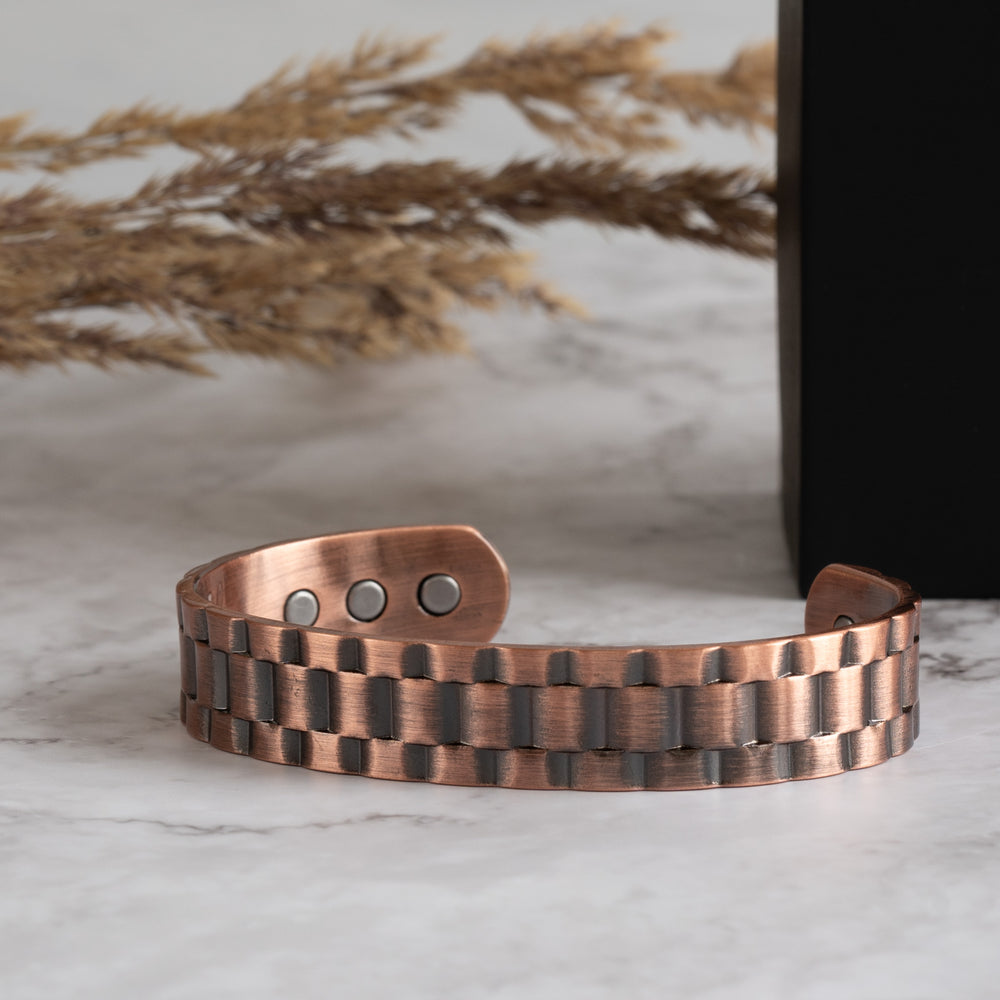 Close-up of the copper bracelet from Magnetic Mobility, showcasing its textured pattern and Neodymium magnets embedded in the back, set on a marble surface with dried golden grass and a black branded box in the background