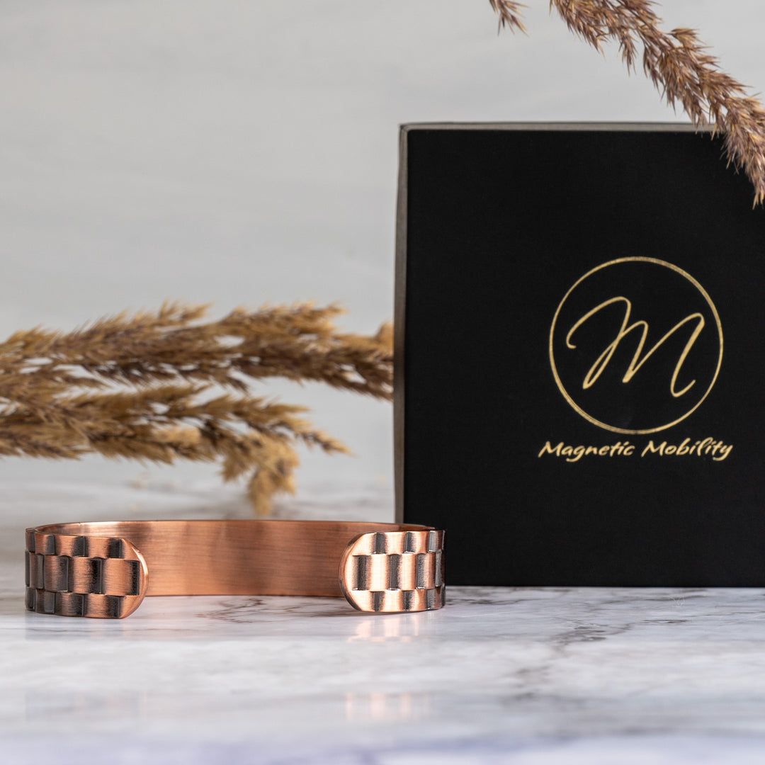 Rear view of the Columbine Copper Bracelet showcasing its open-backed design, positioned in front of a black Magnetic Mobility box on a marble backdrop with a golden dried grass detail