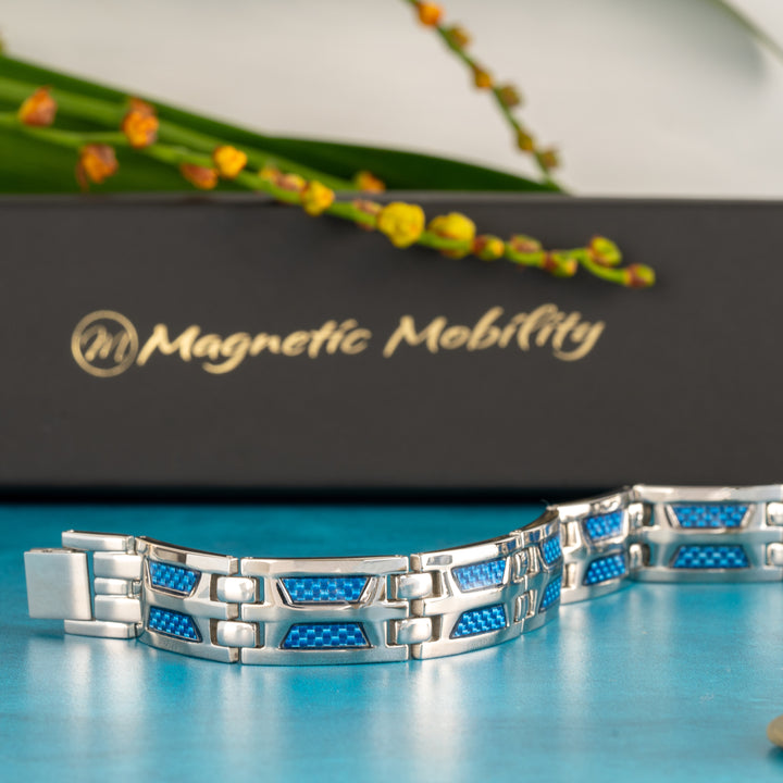 The Yarrow Star 4in1 Magnetic Bracelet by Magnetic Mobility is artfully displayed, half open to reveal the intricate link design with blue carbon fiber inserts. The image captures the bracelet's quality clasp and therapeutic magnets, set against a blue background with the brand's elegant black and gold box and a graceful green plant in soft focus, perfect for shoppers seeking both style and wellness benefits