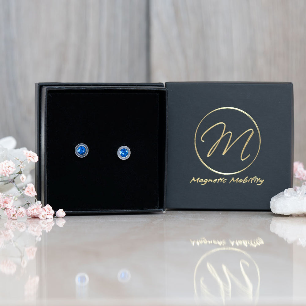 December birthstone magnetic stud earrings with Swarovski Blue Zircon, showcased next to a blue gemstone and presented against a black gift box with Magnetic Mobility logo.