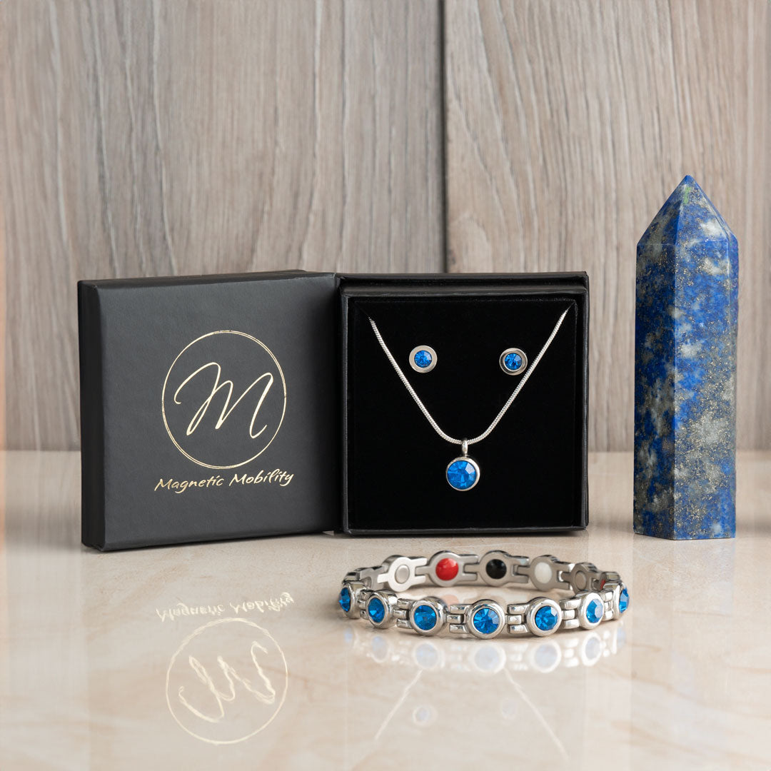 December birthstone gift set including a 4in1 magnetic bracelet, a magnetic necklace, and magnetic stud earrings with Swarovski Blue Zircon, showcased in a black presentation box with Magnetic Mobility branding and a blue crystal obelisk.