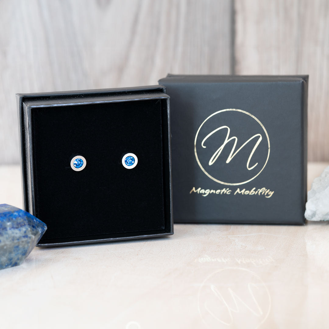 Pair of December birthstone magnetic stud earrings set with vibrant Swarovski Blue Zircon, presented in a black gift box with Magnetic Mobility logo, accompanied by a piece of blue gemstone for an elegant display.