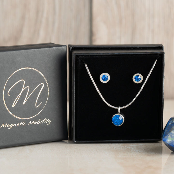 Complete December birthstone gift set featuring a magnetic necklace and matching magnetic stud earrings with Swarovski Blue Zircon, presented in a luxurious black box with Magnetic Mobility logo, displayed with a polished blue gemstone