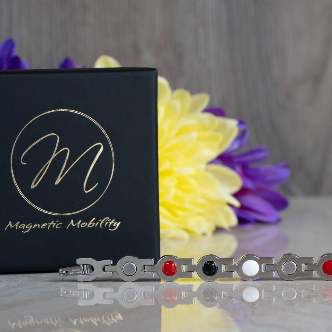 View of the back of a 4in1 Magnetic bracelet showing the 4 elements, magnets, FIR element, Germanimum and Negative ions. There is a black gift box in the background and yellow and purple flowers. 