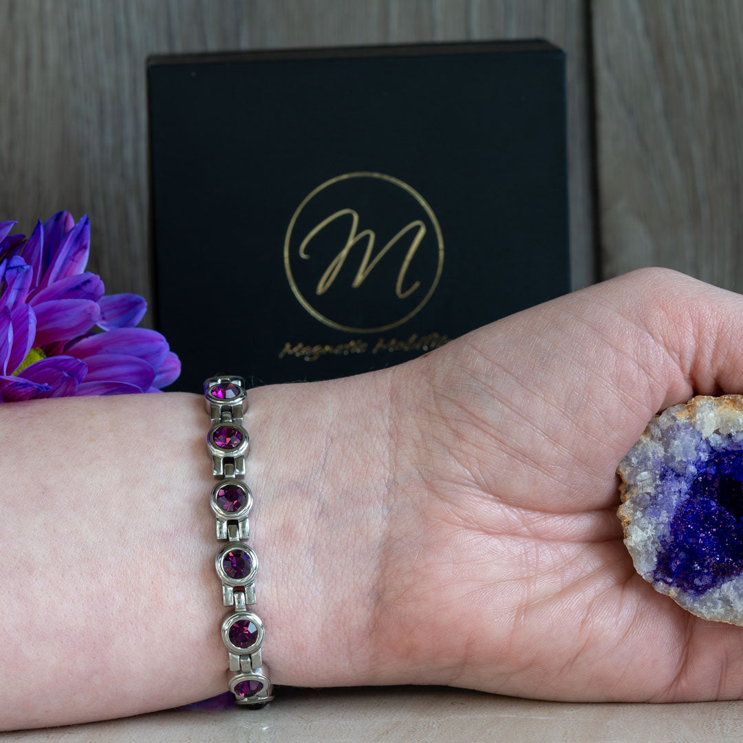 a 4in1 Magnetic bracelet with February birthstone Swarovski Amethyst crystals on a wrist. The hand is holding an amethyst geode and there is a black gift box in the background. 