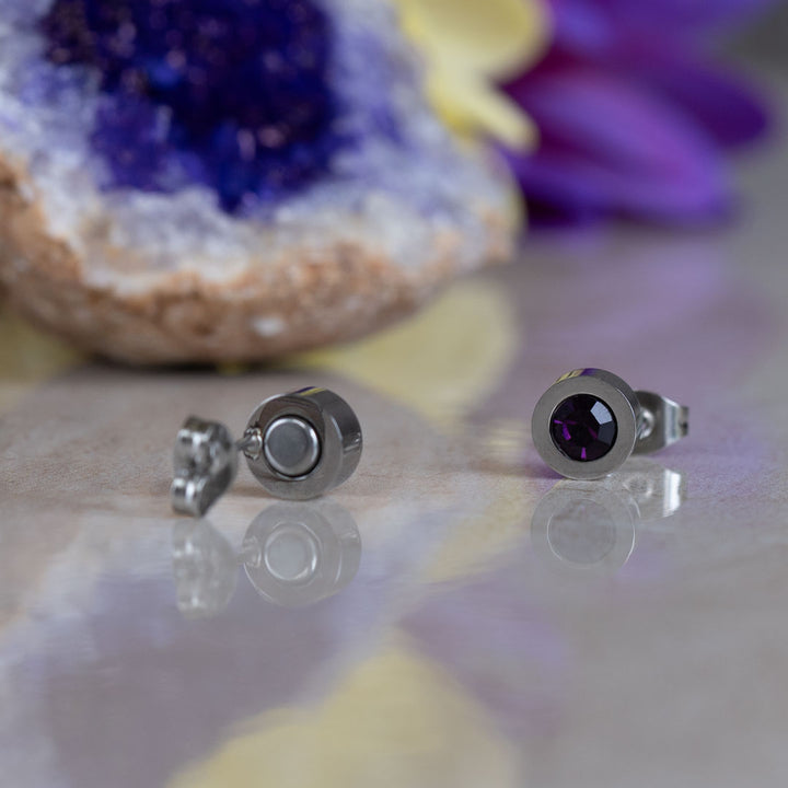 a pair of February Birthstone magnetic earrings with Neodynimum magnets in the back. One bracelet is facing foward shoing the crystal, the second earring is facing backwards showing the magnet. There is an amethyst geode in the background