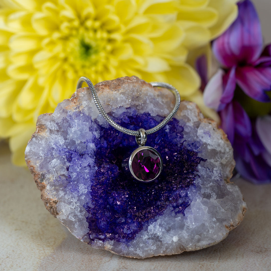 a February birthstone necklace with one Swarovski Amethyst crystal and a neo-dynimum magnet in the back. The Necklace is lying on an amethyst geode with yellow and purple flowers in the background. 