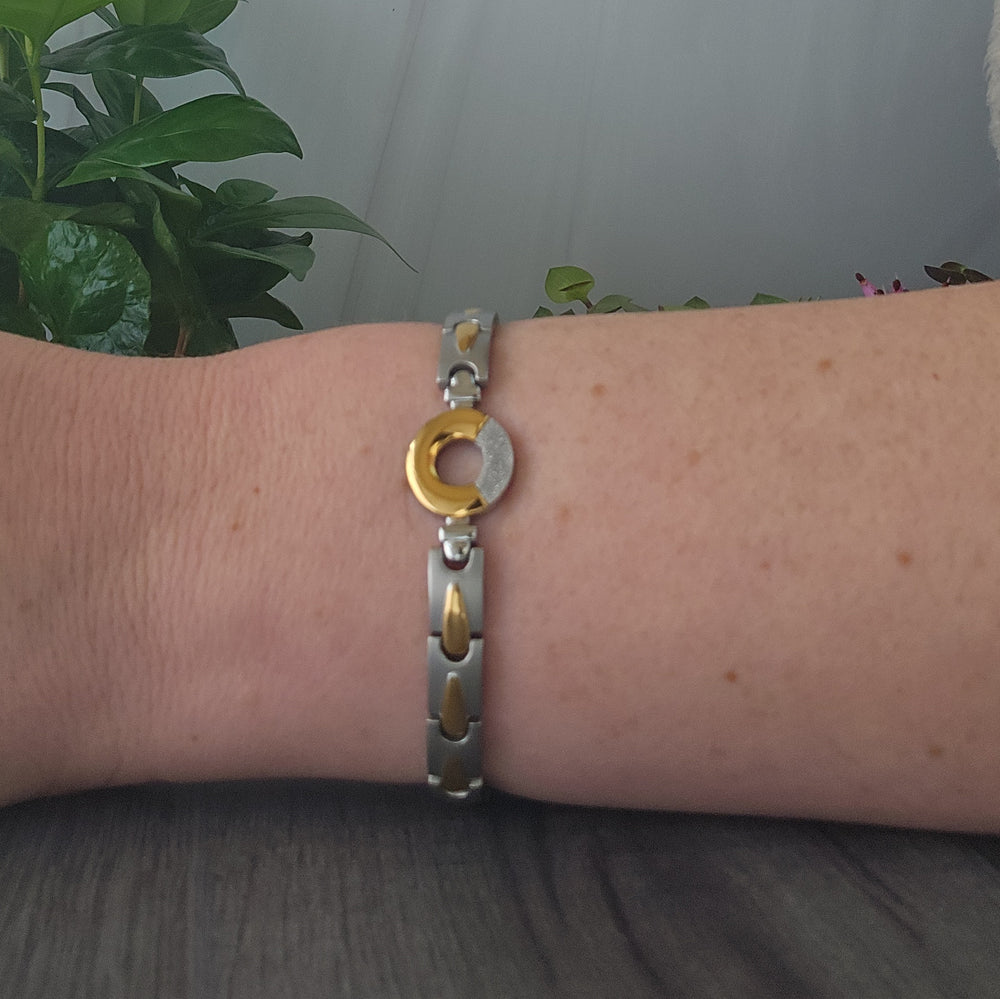 A person's wrist is adorned with the Sorrel Full Moon 4in1 Magnetic Bracelet from Magnetic Mobility, showcasing a sleek design with silver and golden links and a distinctive circular gold center. The background is softly blurred, highlighting the bracelet and a lush green plant, suggesting both style and a connection to nature