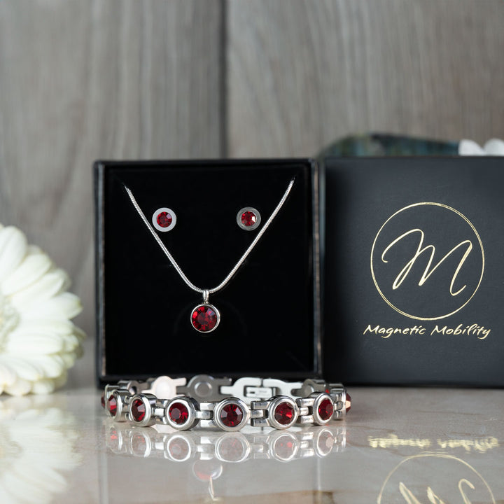 The complete Magnetic Mobility July Birthstone magnetic gift set featuring a necklace, earrings, and bracelet with Swarovski Ruby crystals, presented in luxurious black packaging with a white flower backdrop. Perfect for July birthday gifts, combining elegance and health benefits.