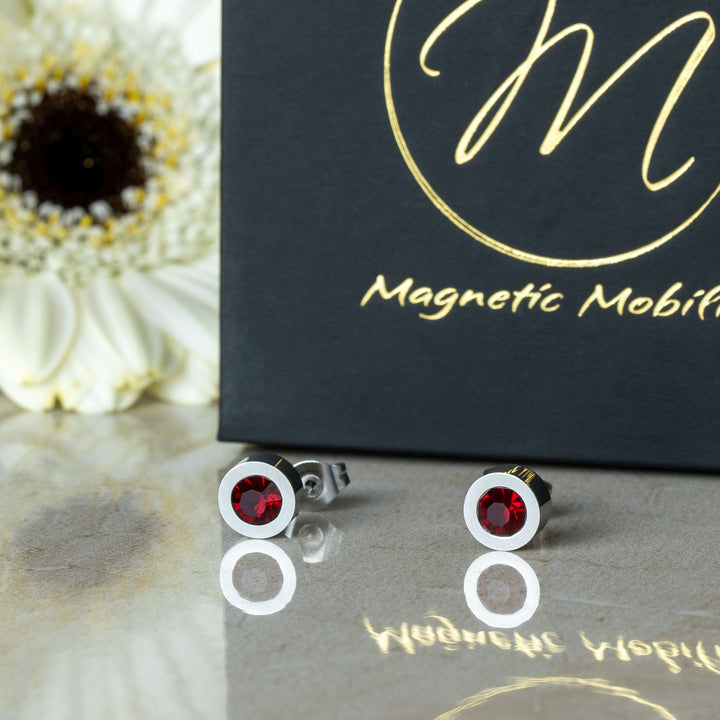 Front view of the July Birthstone magnetic earrings featuring radiant Swarovski Ruby crystals, displayed on a reflective surface with a white flower and black box backdrop. The earrings are designed for elegance and therapeutic benefits, perfect for July birthdays.