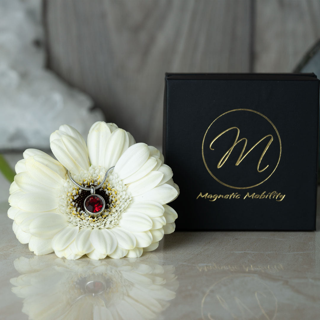 The July Birthstone magnetic necklace with a vibrant Swarovski Ruby pendant, beautifully displayed on a white flower with a black Magnetic Mobility gift box in the background. Ideal for celebrating July birthdays with style and grace.