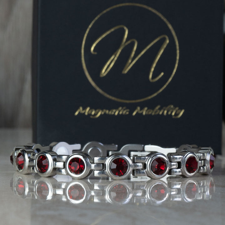 Close-up of the July Birthstone magnetic bracelet adorned with striking Swarovski Ruby crystals, displayed on a reflective surface with a black gift box background. The bracelet combines the fiery beauty of rubies with therapeutic benefits, perfect for July birthday gifts.