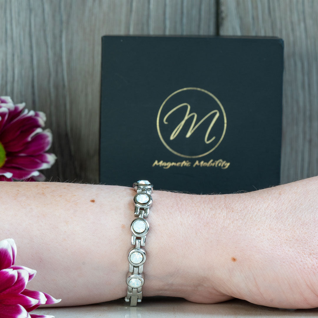 The June Birthstone magnetic bracelet featuring Swarovski Pearls is shown on a wrist, highlighting its sophisticated design and comfortable fit. The backdrop includes a dark Magnetic Mobility box and a pink flower, emphasizing the bracelet's elegance and appeal.