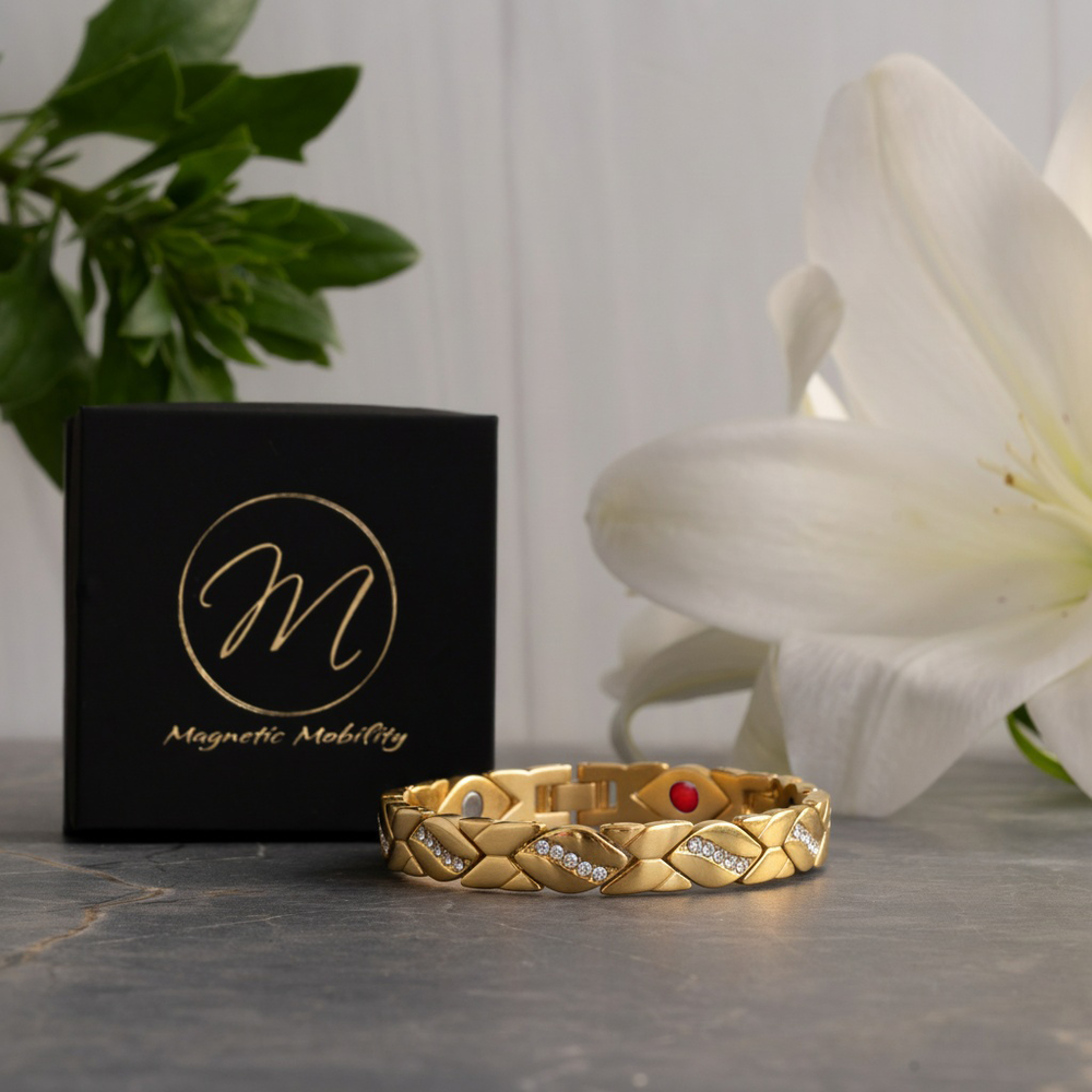 Gold magnetic bracelet with decorative elements on a gray surface next to a black box with 'Magnetic Mobility' branding, and a white flower in the background.