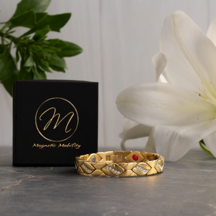 Gold magnetic bracelet with decorative elements on a gray surface next to a black box with 'Magnetic Mobility' branding, and a white flower in the background.