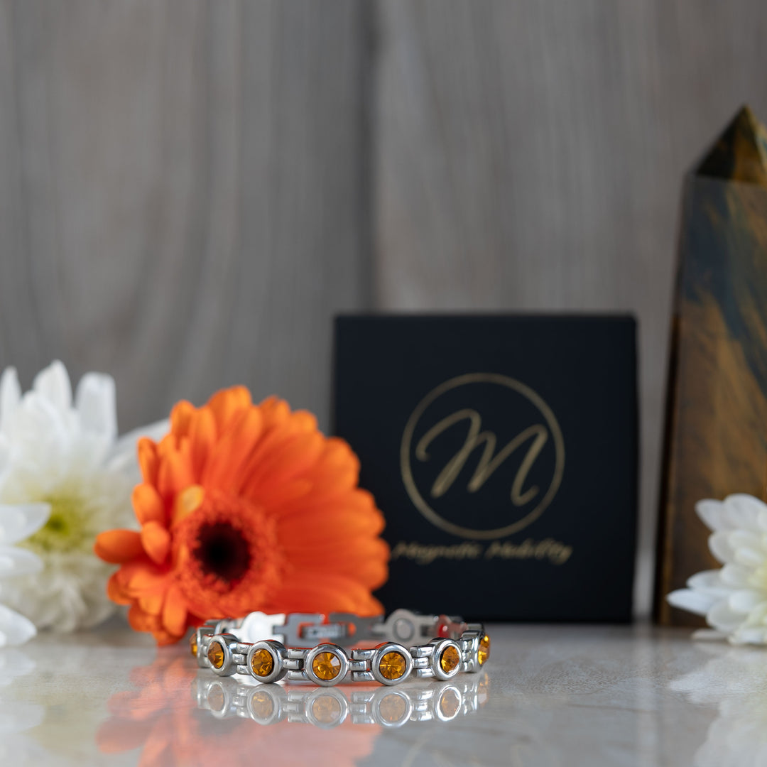 A striking 4in1 magnetic bracelet with multiple citrine stones, symbolizing the November birthstone. The bracelet is set against a backdrop of white and orange flowers, complementing the bracelet’s warm, golden tones. The Magnetic Mobility logo can be seen on a black box in the background.