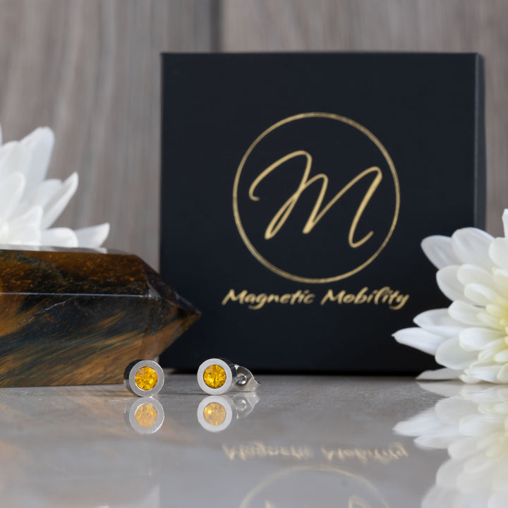 A front view of the magnetic stud earrings with citrine stones, representing the November birthstone. The earrings are placed against a dark background, with a polished stone to the left and white flowers in the backdrop. The Magnetic Mobility logo is visible in the background.