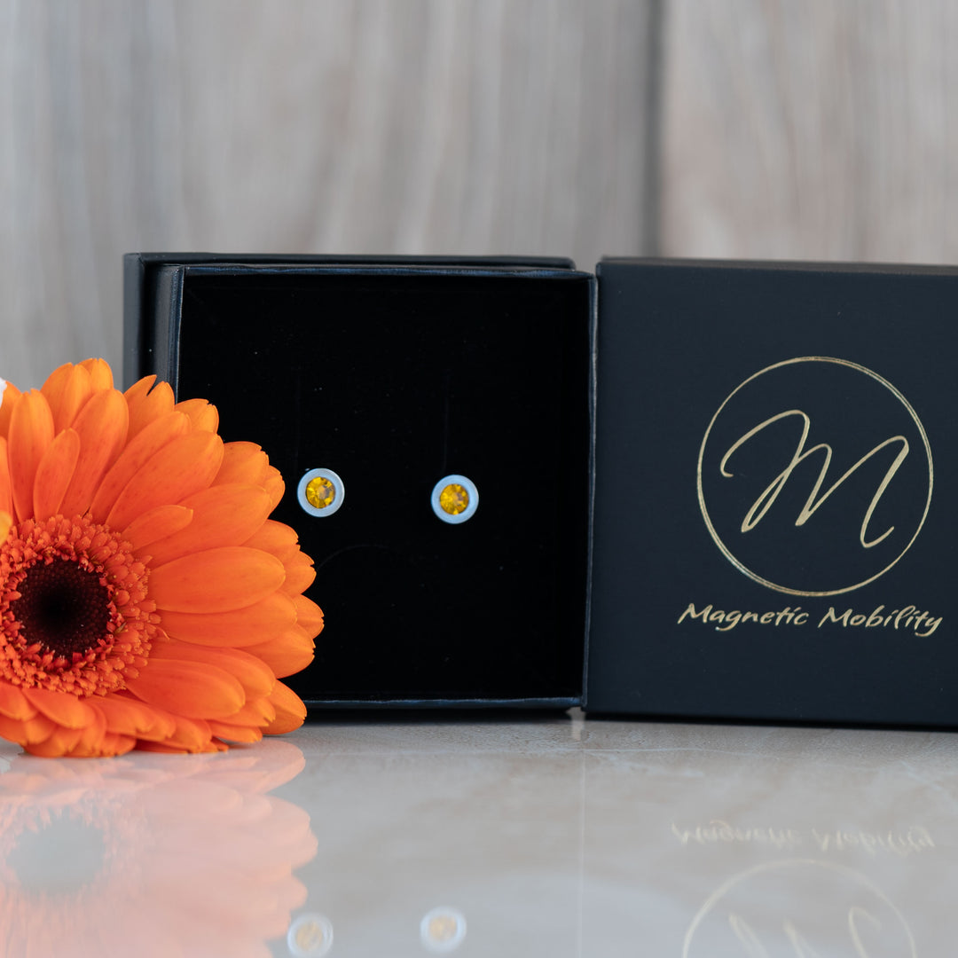 A close-up of the November birthstone earrings featuring vibrant citrine stones. The earrings are displayed in a black box with an orange flower beside them, highlighting their bright, sunny yellow hue. The Magnetic Mobility logo is visible on the box.