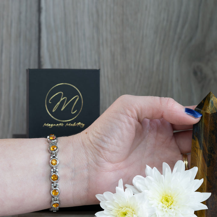 The November birthstone magnetic bracelet, adorned with citrine stones, is elegantly displayed on a wrist. The warm yellow stones are contrasted against the wearer’s skin, with a background featuring the Magnetic Mobility logo and white flowers.