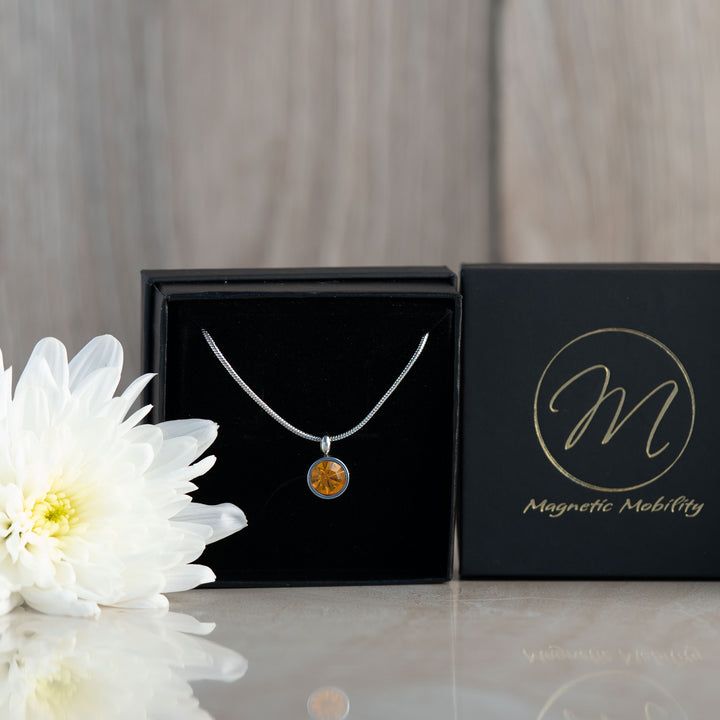 A close-up of the November birthstone necklace featuring a round citrine pendant. The necklace is elegantly displayed in a black box, with a white flower beside it, highlighting the pendant's warm, golden-yellow hue and the Magnetic Mobility logo in the background.