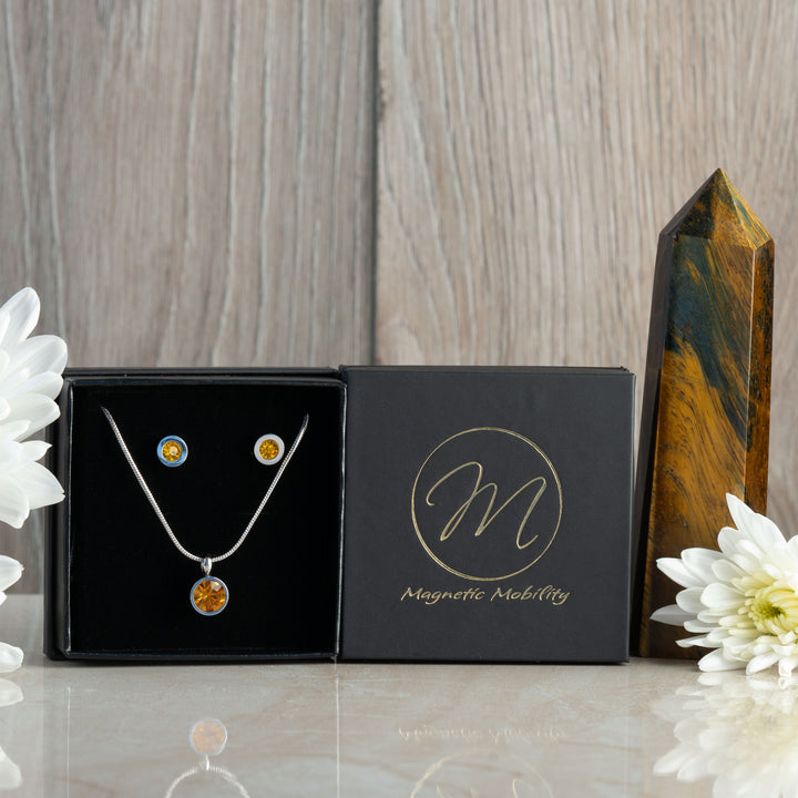 Close-up of the November birthstone necklace and earrings set, featuring bright citrine stones. The jewelry is elegantly displayed in a black box with an orange flower nearby, emphasizing the citrine's vibrant color and the Magnetic Mobility logo on the box.