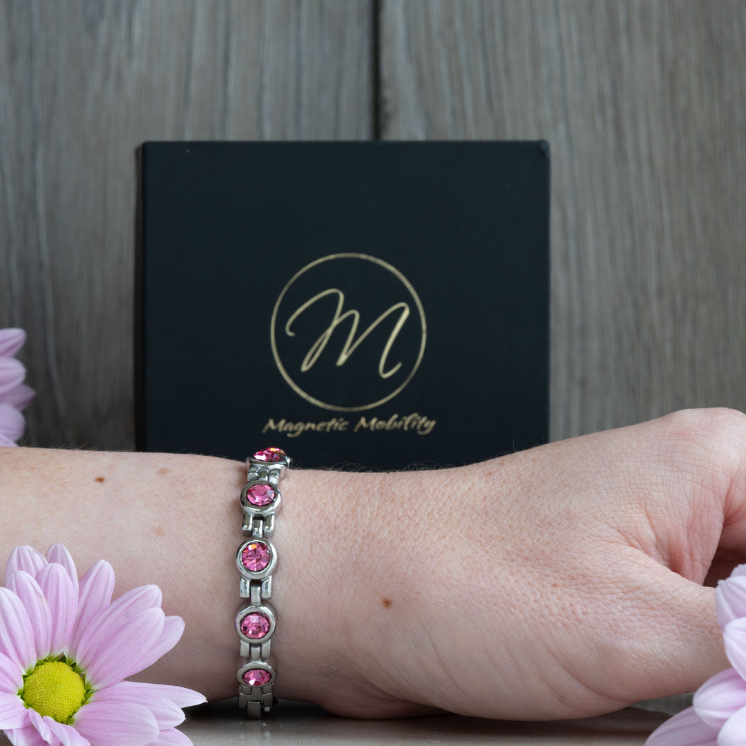 Side view of a pink tourmaline magnetic bracelet from Magnetic Mobility, worn on a woman’s wrist. The bracelet features multiple sparkling pink crystals set in a stylish silver band, accompanied by pink flowers and the brand’s packaging.