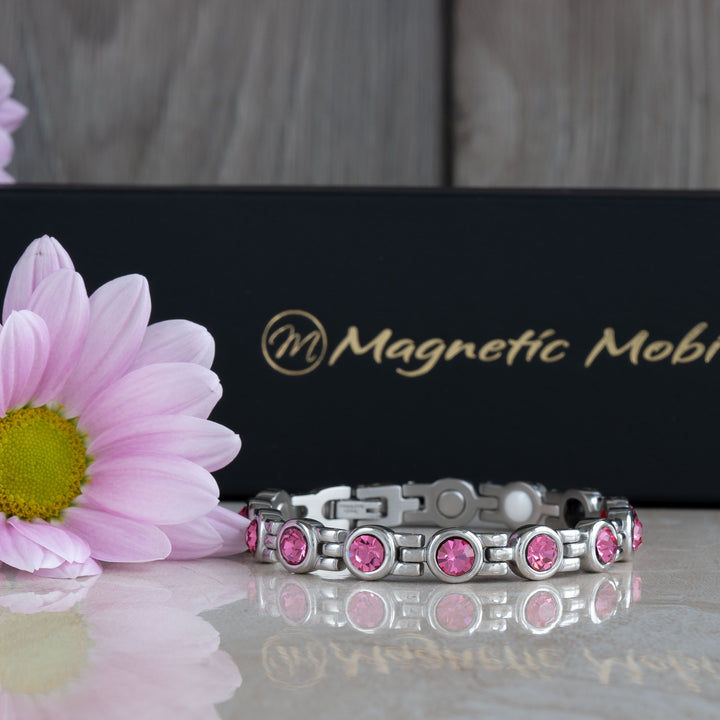 Full view of a pink tourmaline magnetic bracelet from Magnetic Mobility, laid flat to showcase its design. The bracelet features a series of pink crystals set in a silver band, accompanied by a pink flower and the brand’s elegant black packaging.