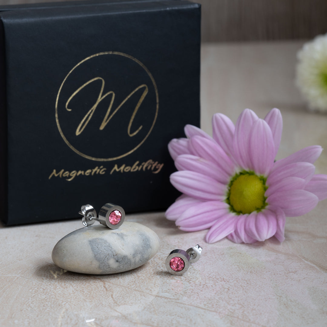 Close-up image of pink tourmaline magnetic stud earrings from Magnetic Mobility, highlighting the sparkling pink crystals set in a silver-toned base. The earrings are displayed with a pink flower and the brand’s packaging.