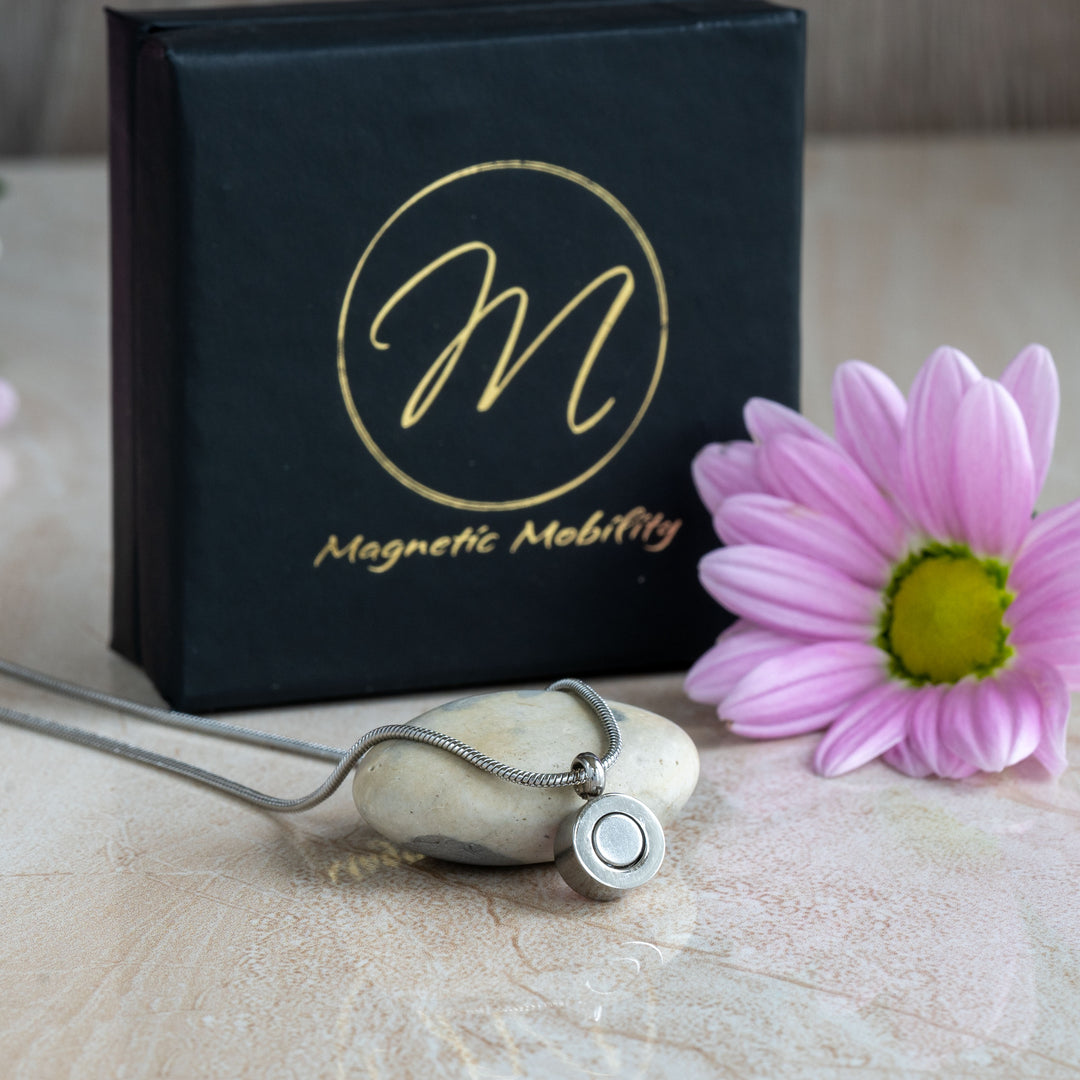 Back view of a pink tourmaline magnetic necklace from Magnetic Mobility, showing the sleek pendant and adjustable snake chain. The necklace is placed beside a pink flower and the brand’s black gift box.